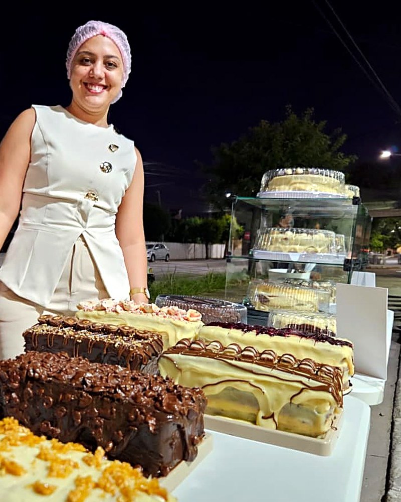 A Leyde sorrindo atrás da mesa de tortas no Festival de Fatias, na praça, à noite