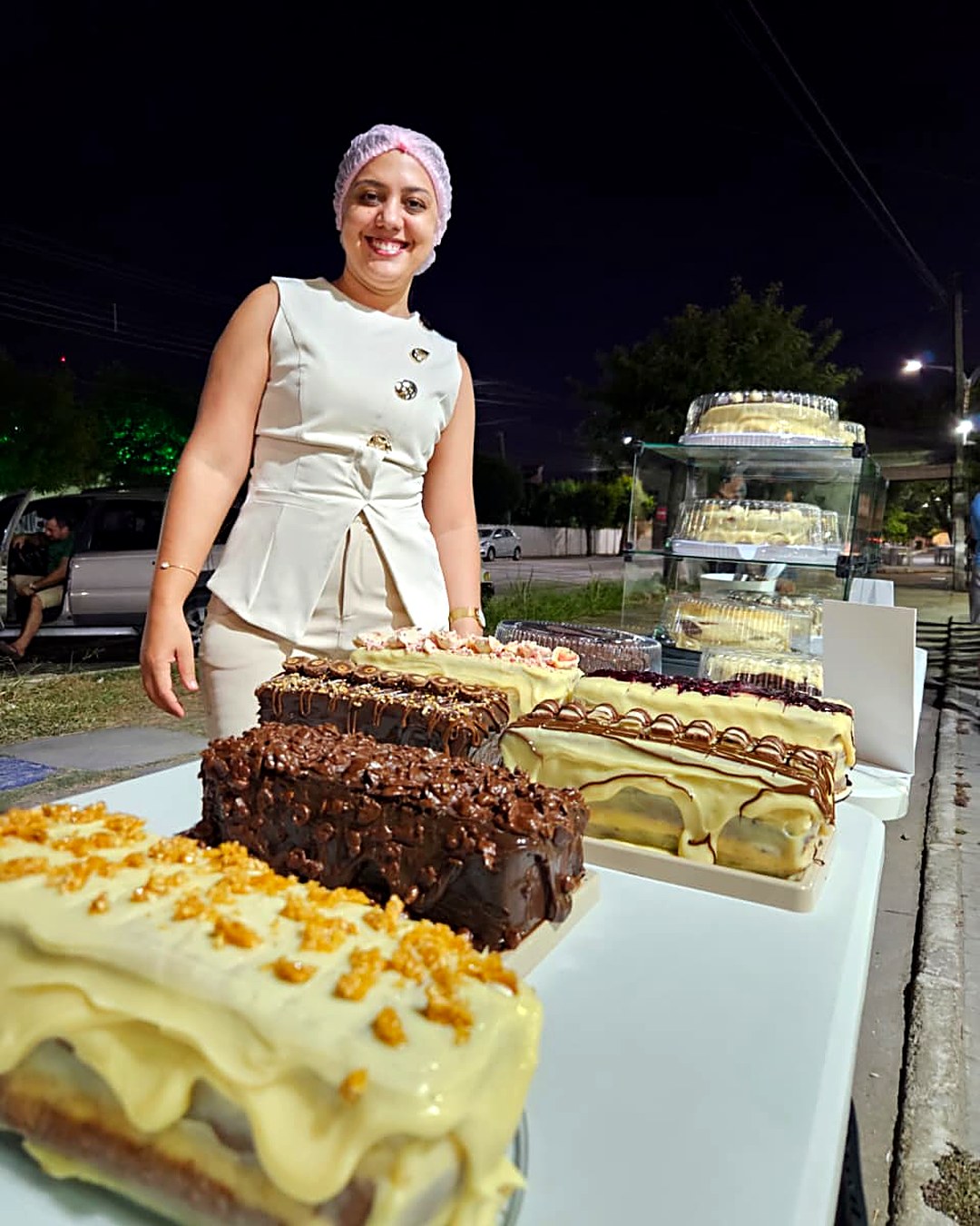 Leyde sorrindo atrás da mesa do Festival, com os bolos inteiros à frente e a vitrine de vidro ao fundo, na Praça da Rodoviária